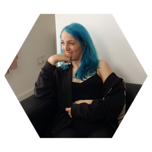 Liz sits on a sofa in black crop top and sweatpants, has blue hair, and is smiling with her chin resting on her hand, looking off-camera